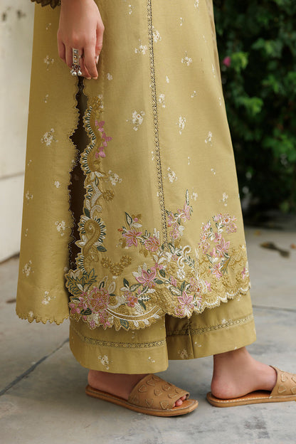 A close-up of a person wearing a mustard yellow embroidered kurta and matching wide-leg trousers, with floral embroidery concentrated along the hem and side slit. The person is wearing tan slide sandals, standing outdoors on a stone floor with greenery in the background.