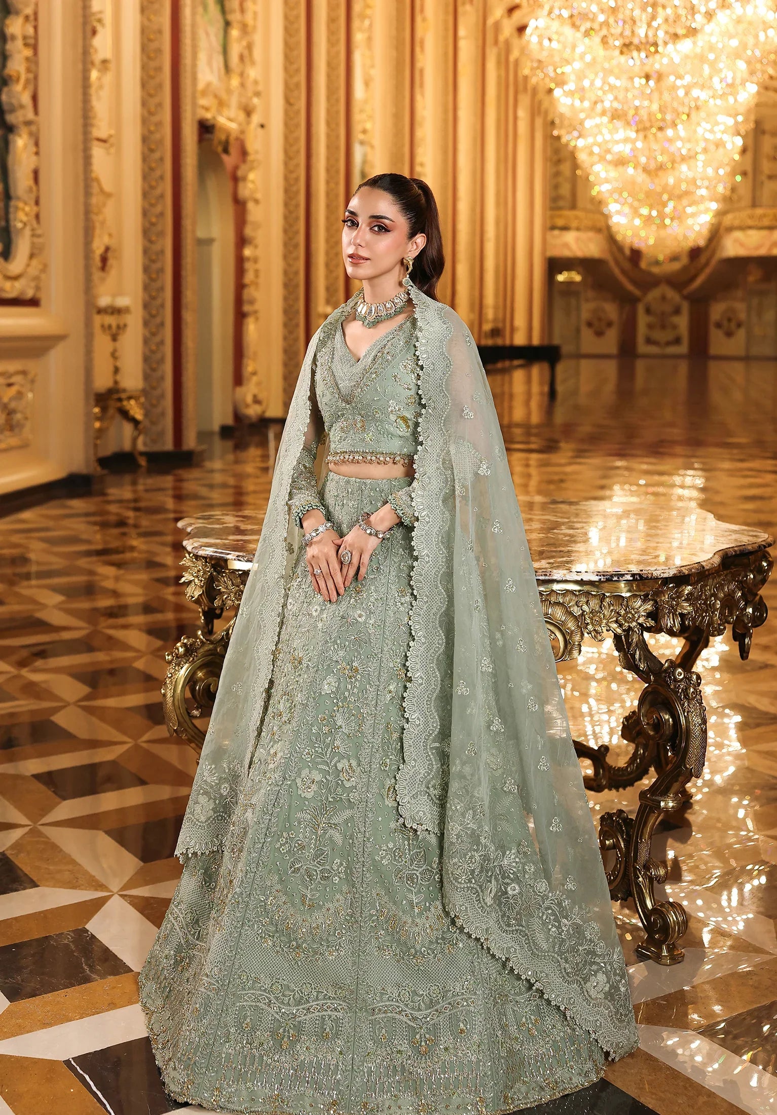 Woman in a traditional green outfit with a long train standing in an opulent room with chandeliers.