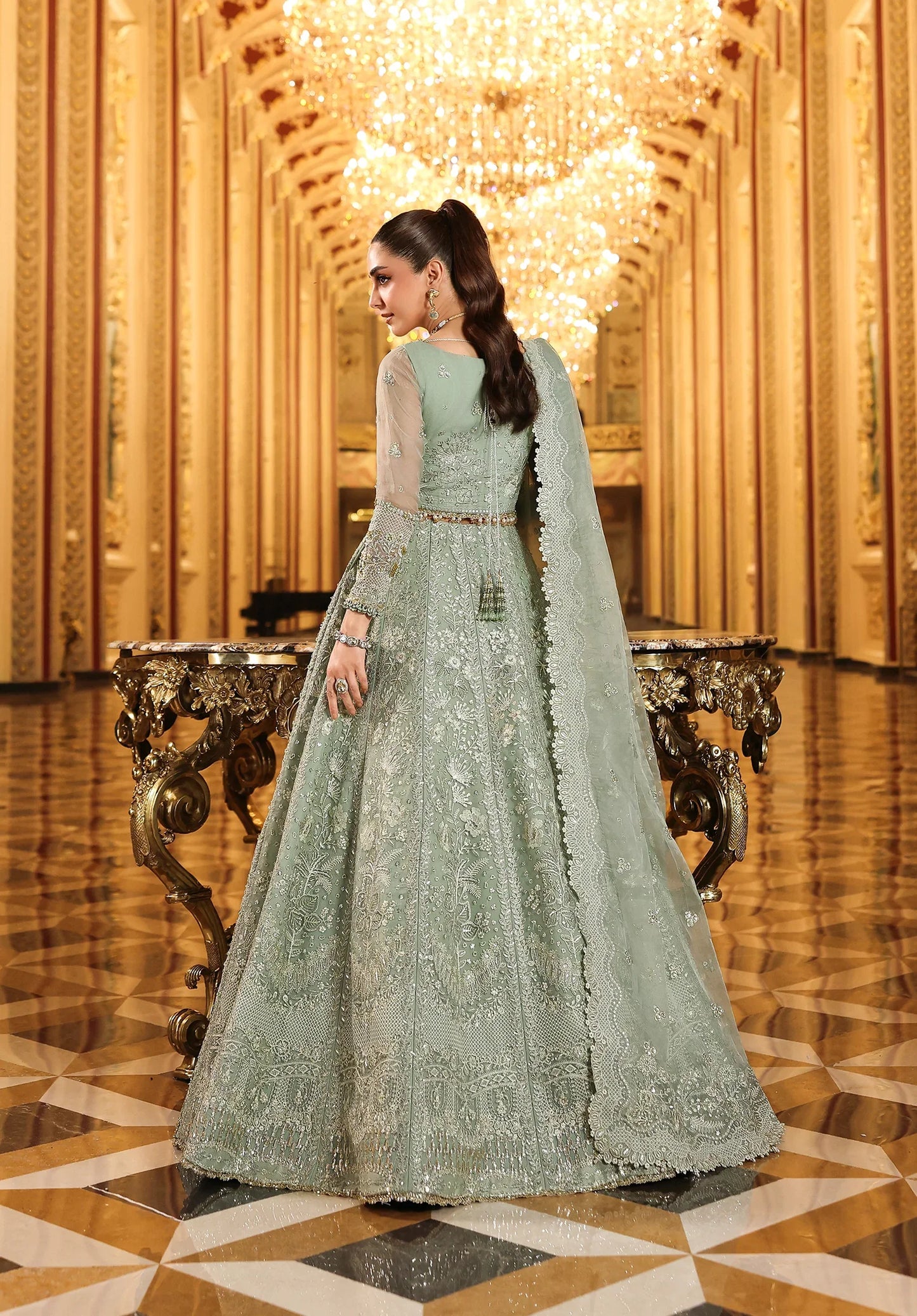 Woman in a light green embroidered dress standing in an ornate hall with gold decorations.