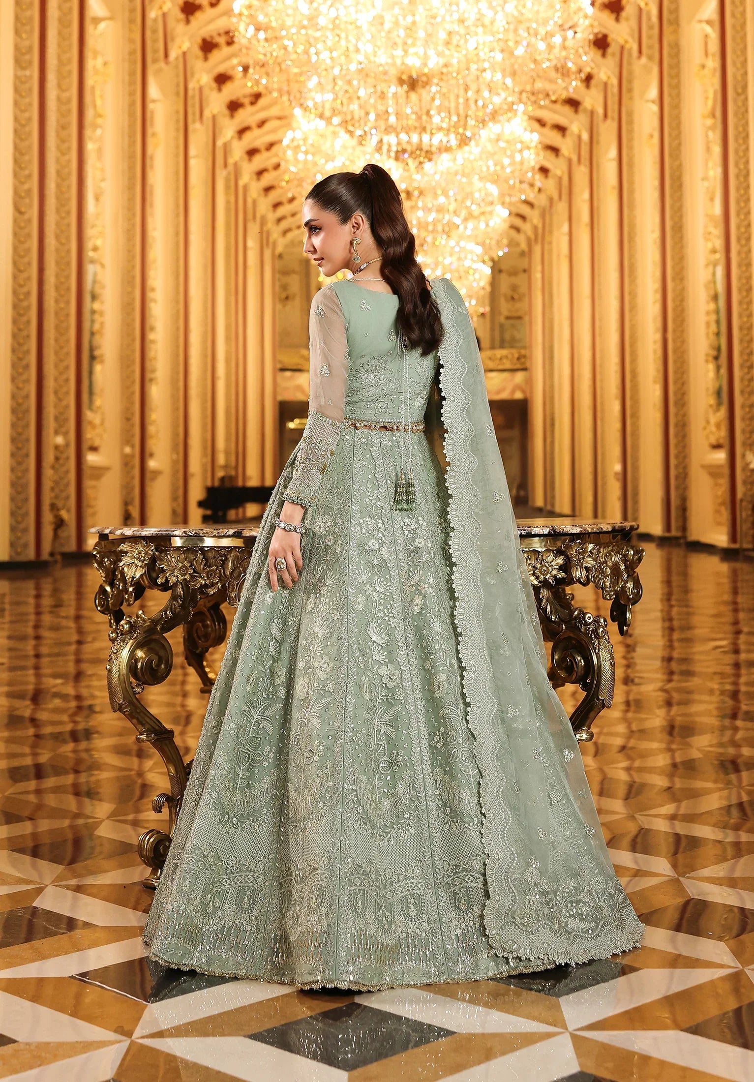 Woman in a light green embroidered dress standing in an ornate hall with gold decorations.