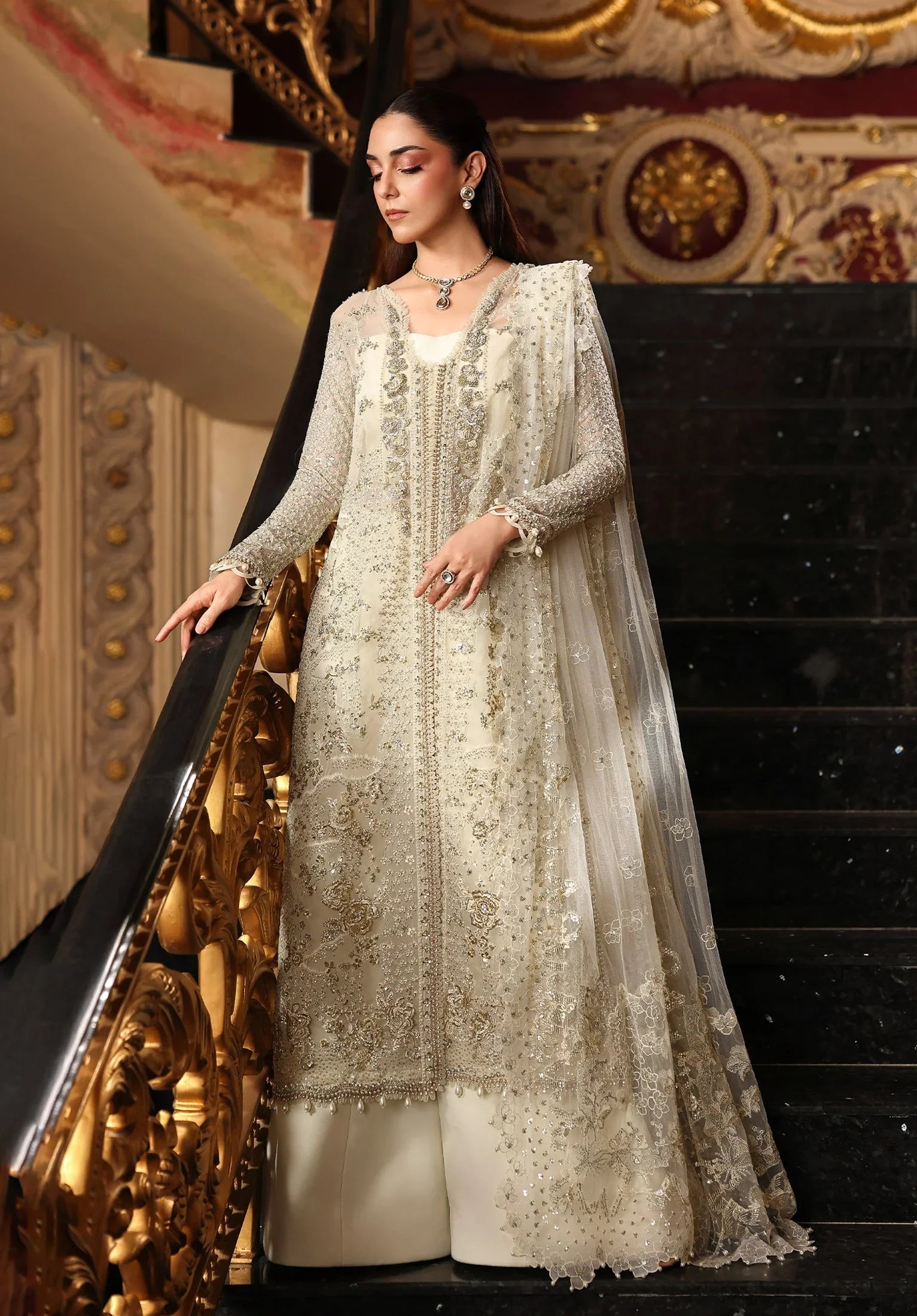 Woman in a traditional embroidered outfit standing on a staircase with ornate decorations with Elaf Stardom Handwork.